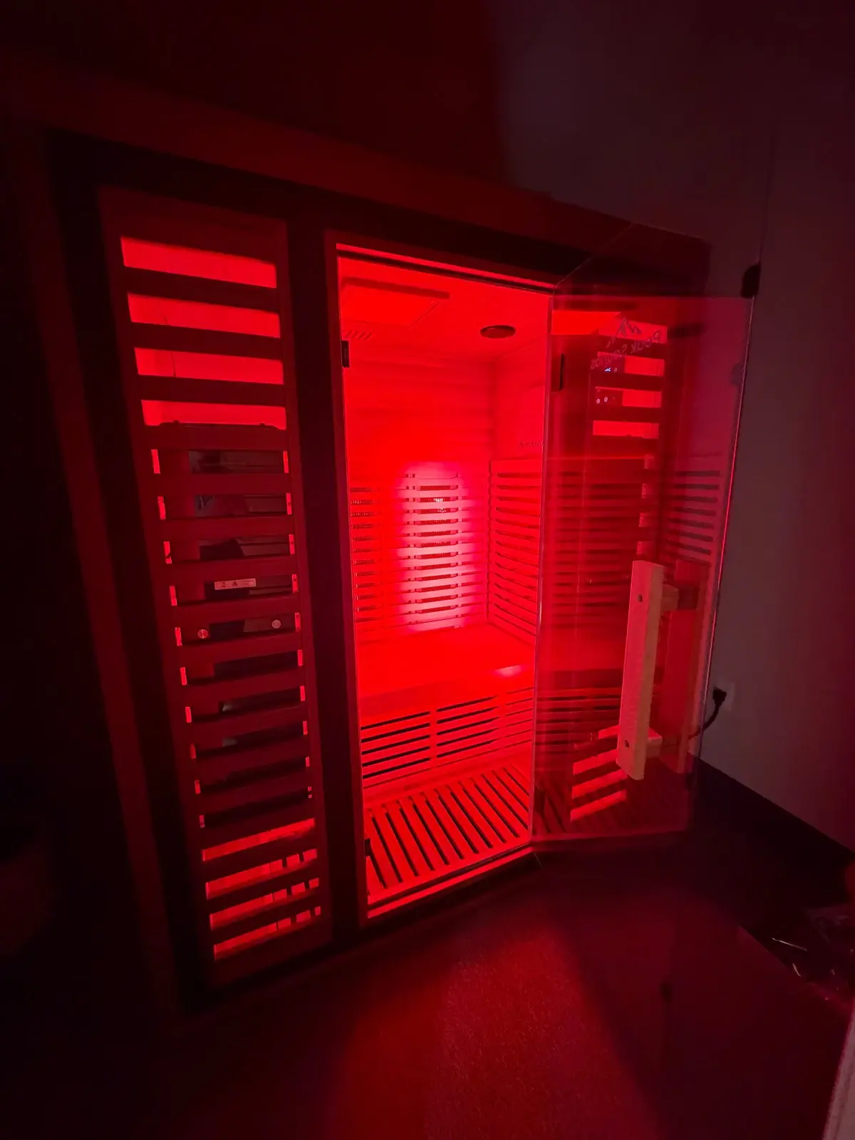 Infrared sauna with its glass door open, showing the wooden bench and red light glowing inside